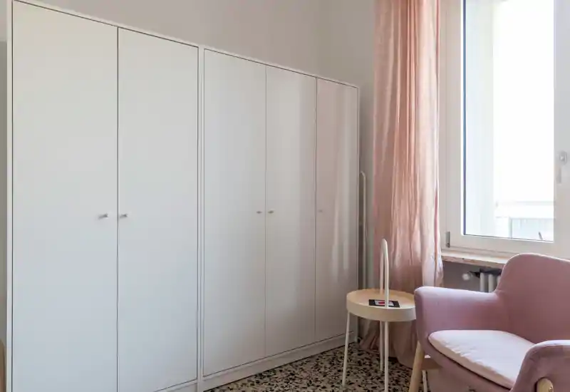Minimal bedroom corner with a large white wardrobe, soft pink armchair, side table and a bright window — a calm, functional dressing area with gentle natural light.