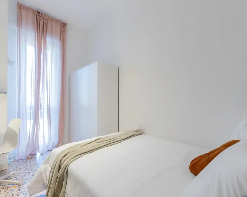 Camera da letto luminosa e minimale con letto matrimoniale rifatto, coperta morbida e una grande finestra con tende rosa leggere che lasciano entrare luce naturale.