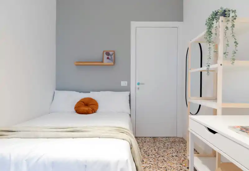 Camera da letto luminosa e minimale con letto matrimoniale ben rifatto, cuscino accentato caldo e mensola in legno sopra la testata — composizione pulita ideale per un annuncio.