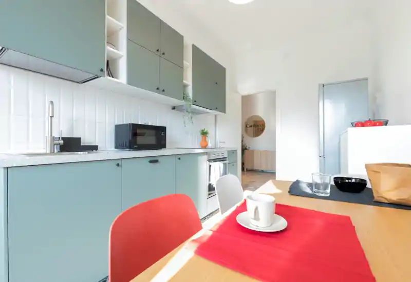 Bright, modern kitchen with muted green cabinets, white tiled backsplash and a wooden dining table set with a cup and placemat — a clean, welcoming space ideal for short-term rentals.