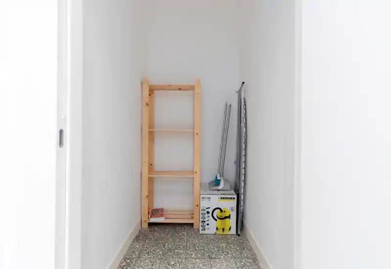 Small entrance or storage alcove with a wooden shelving unit, cleaning tools and an ironing board on patterned tile flooring. Bright, minimal and functional space suitable for storage.