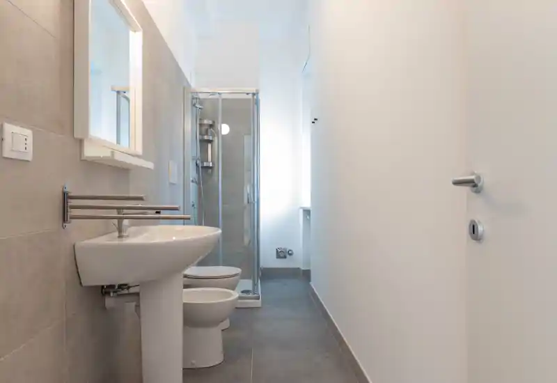 Compact modern bathroom with a pedestal sink, toilet and a corner glass shower. Neutral grey tiles and bright white walls create a clean, minimalist look.