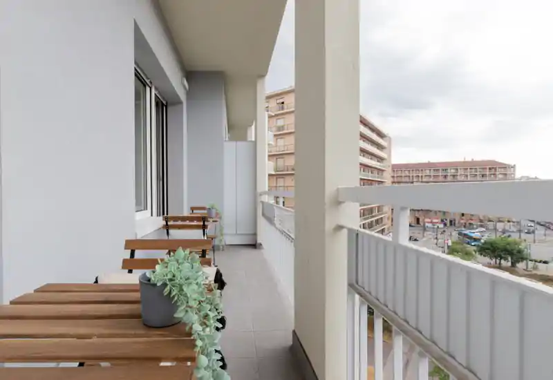 Sunny city-facing balcony with narrow tiled floor, wooden bistro tables and planter greenery — ideal for morning coffee and urban views.