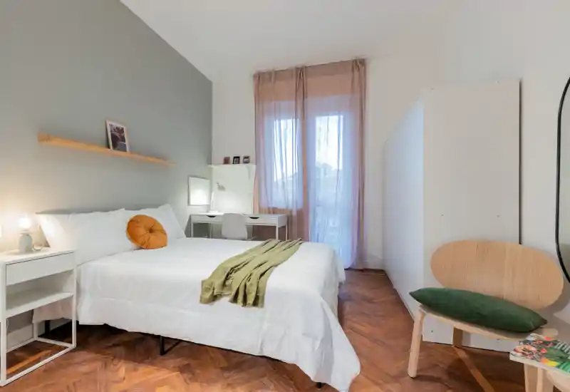 Chambre lumineuse et soignée avec lit double bien fait, table de chevet, petit bureau près de la fenêtre et armoire — tons chauds et parquet en chevrons créant une atmosphère cosy.