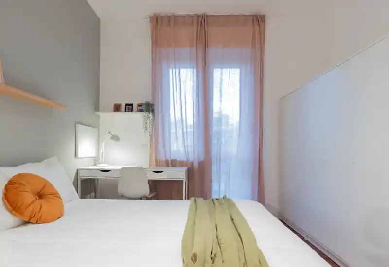 Chambre lumineuse au style doux avec grand lit blanc, coussin accent chaleureux et un petit bureau près de la fenêtre bénéficiant de lumière naturelle.