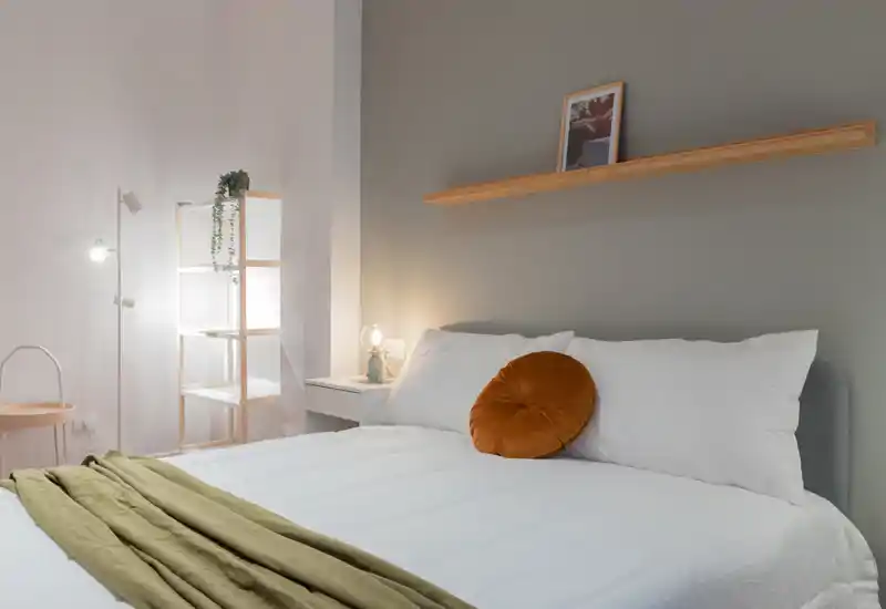 Chambre lumineuse et minimaliste avec lit entièrement visible habillé de linge blanc, coussin rond couleur rouille et éclairage d'ambiance doux. Une étagère simple et une lampe de chevet complètent ce look moderne et chaleureux.
