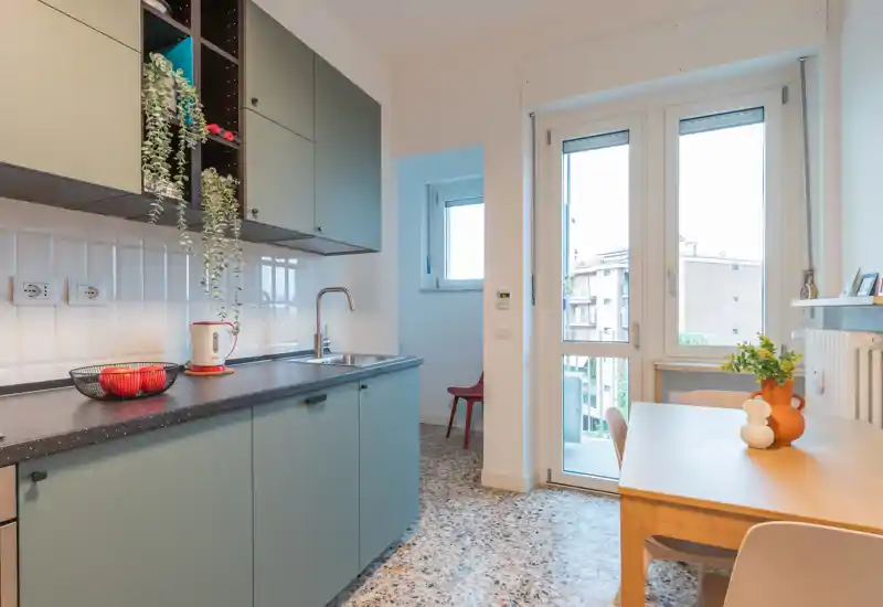 Cuisine moderne et lumineuse avec des placards vert sauge, un plan de travail foncé et une table en bois près d'une grande fenêtre/porte. Plantes et corbeille de fruits apportent une touche chaleureuse.