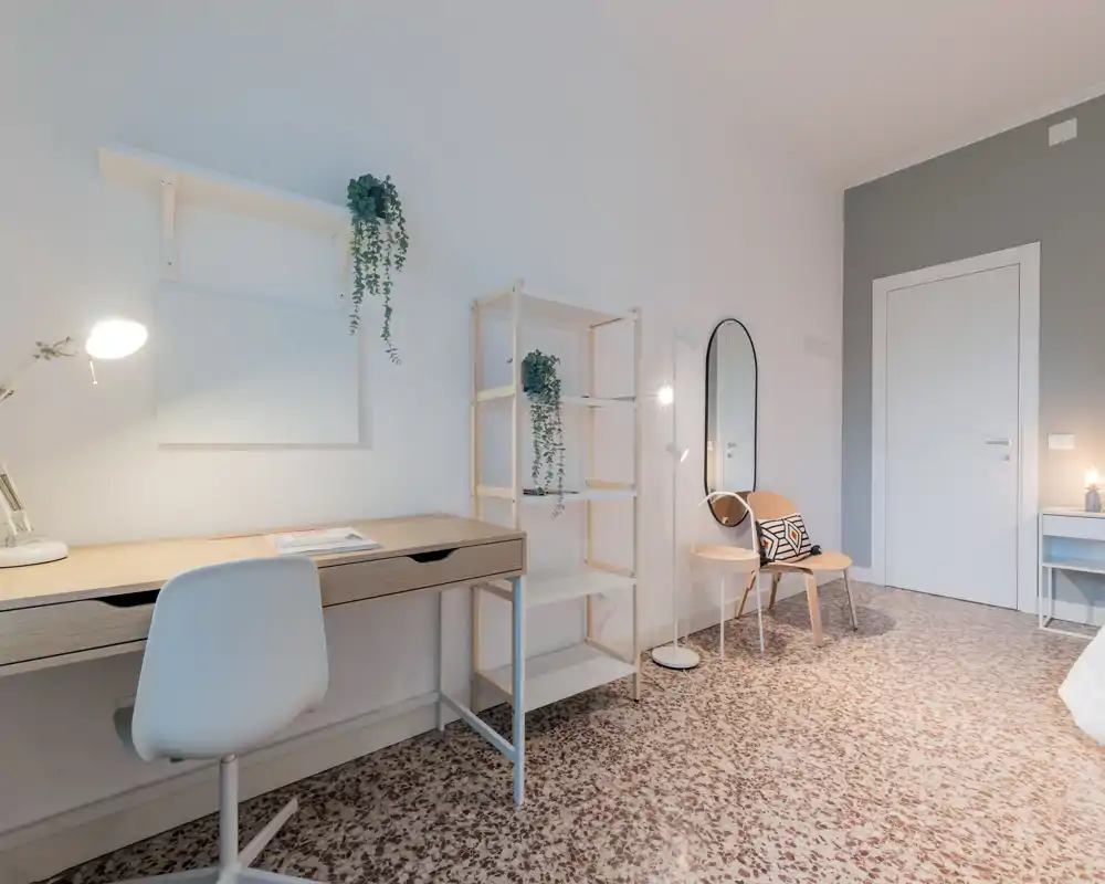 Angolo di camera luminoso e minimal con scrivania in legno, scaffalatura e una zona salotto accogliente. Toni neutri, piante e illuminazione soffusa creano un’atmosfera moderna e rilassante, perfetta per lavoro e riposo.