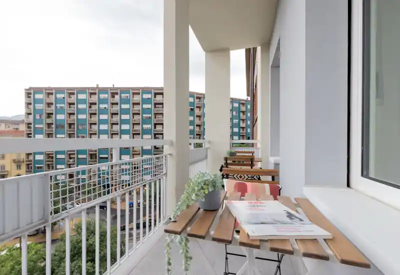 Balcone soleggiato con piccolo tavolo in legno e sedie pieghevoli, piante in vaso e vista sui palazzi vicini — un angolo esterno compatto per caffè o lettura mattutina.