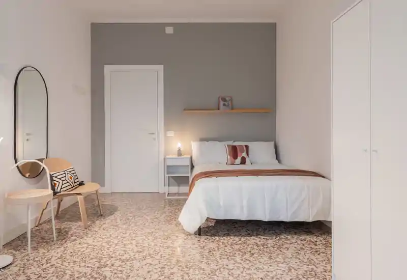 Camera da letto luminosa e minimal con letto matrimoniale centrale, comodino con lampada, cuscini decorativi e palette neutra per un'atmosfera rilassante.