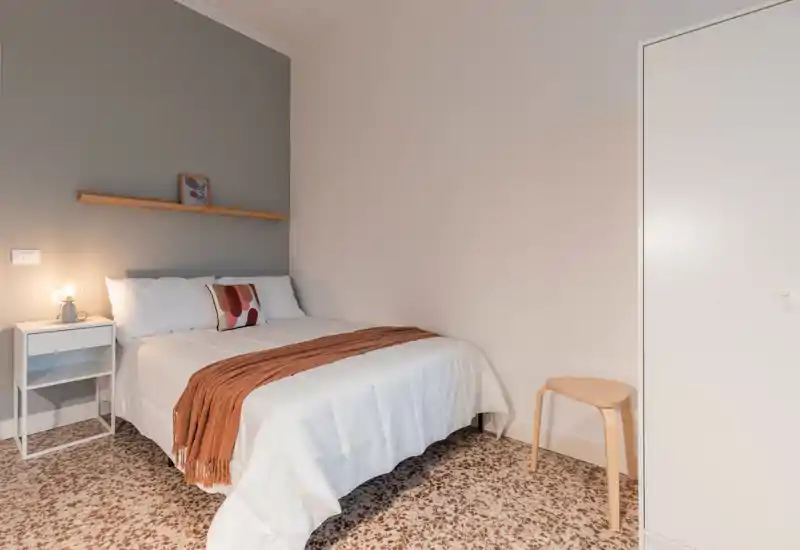 Camera da letto luminosa e curata, con letto bianco, plaid caldo e cuscino decorativo; arredo minimale e comodino con lampada per un ambiente accogliente.