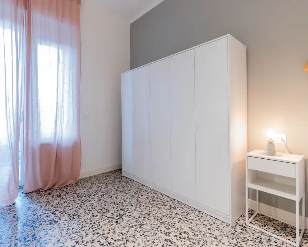 Coin de chambre minimaliste avec une grande armoire blanche, une table de chevet avec lampe et des rideaux roses légers laissant passer la lumière naturelle — propre, moderne et épuré.