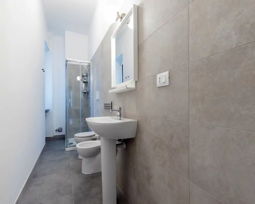 Bagno moderno e compatto con lavabo a colonna, WC, bidet e cabina doccia in vetro. Piastrelle grigie neutre e sanitari bianchi creano un ambiente minimal e facile da mantenere.