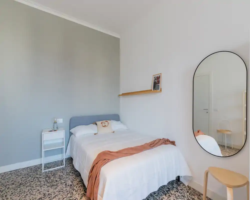 Bright, minimalist bedroom with a double bed, white linens, a warm throw and a large oval mirror. Clean lines and soft natural light create a calm, modern atmosphere ideal for rental photos.