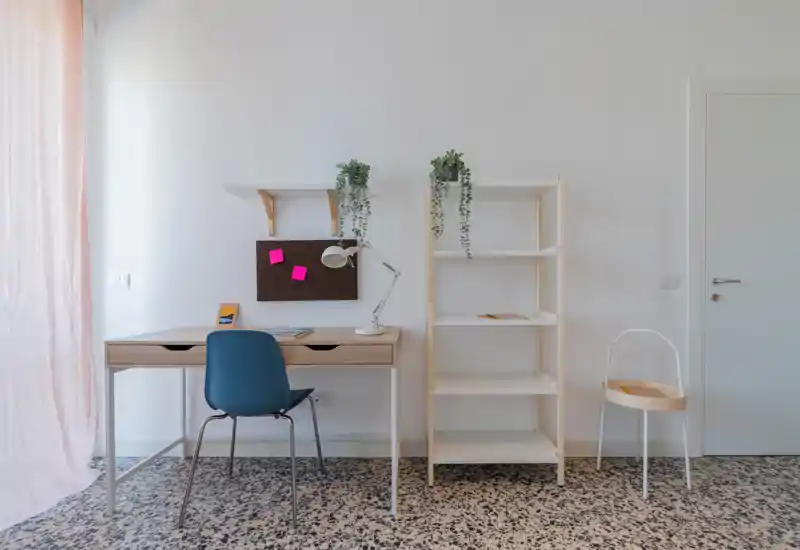 Bright, minimal home office corner with a wooden desk, blue chair, desk lamp and shelving. Soft natural light and hanging plants create a calm, modern workspace.