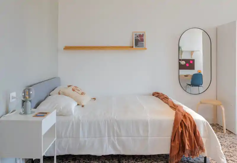 Bright, minimal bedroom with a neatly made double bed, a bedside table with a lamp, decorative cushion and a warm throw. A tall oval mirror and simple shelving complete the clean, contemporary look.