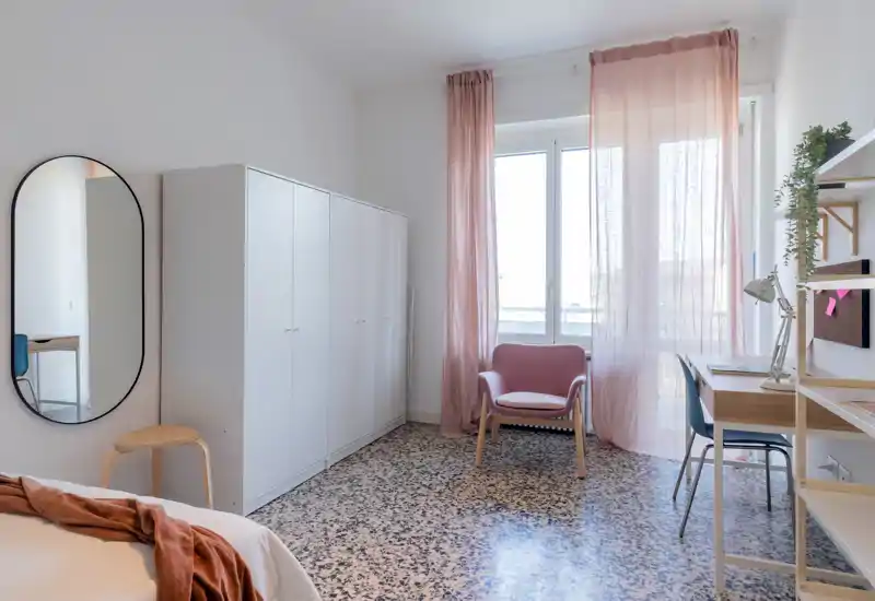 Bright, stylish bedroom with a white wardrobe, a comfortable armchair by the window and a light wooden desk — soft pink curtains add warmth and natural light.