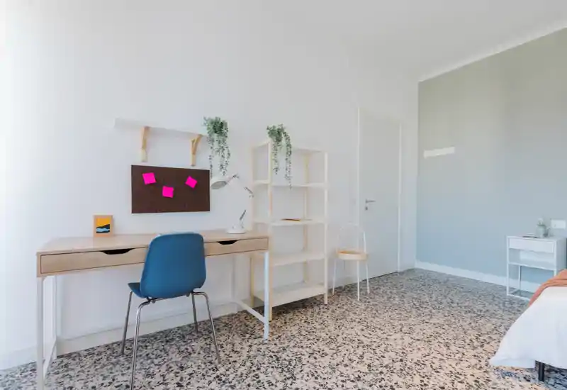 Bright, minimal bedroom with a study corner: a wooden desk, blue chair, shelving and a partially visible bed at the right edge. Clean terrazzo floor, hanging plants and neutral walls create a calm, modern atmosphere.