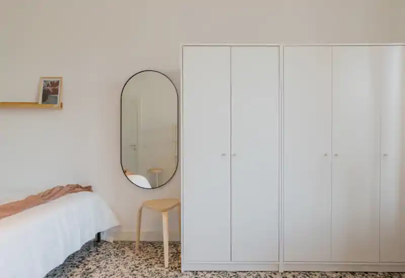 Minimal, bright bedroom with a white wardrobe and a neatly made bed on the left, accented by a soft throw and an oval wall mirror that adds depth.