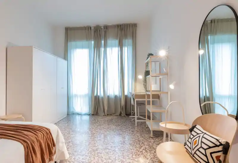 Chambre lumineuse et aérée avec grandes fenêtres voilées, lit bien fait avec plaid chaud, armoire et coin assise avec miroir — style minimal et moderne idéal pour une annonce.