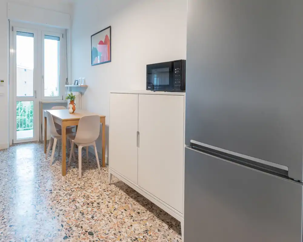 Coin cuisine moderne et compact avec une petite table en bois, quatre chaises, réfrigérateur et micro-ondes; belle lumière naturelle venant de la porte/fenêtre et décoration minimale soignée.