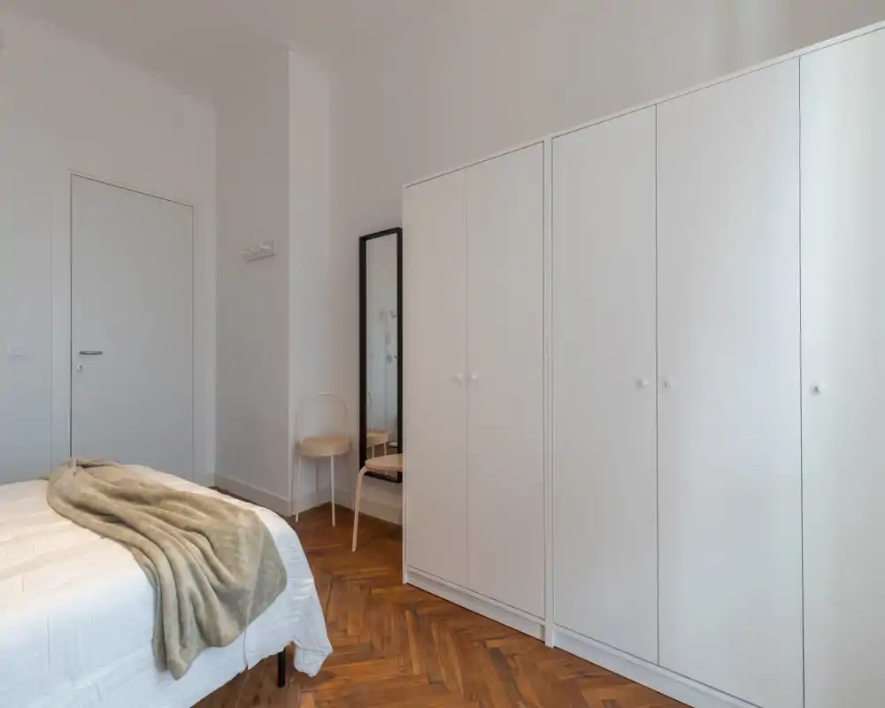 Dormitorio luminoso y minimalista con armario blanco, suelo de parquet y cama bien hecha con una manta suave; espejo y silla añaden funcionalidad.