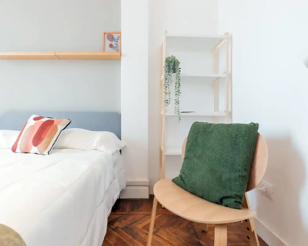 Dormitorio luminoso y minimalista con cama bien hecha, cojín decorativo y una silla de madera que crea un rincón acogedor.