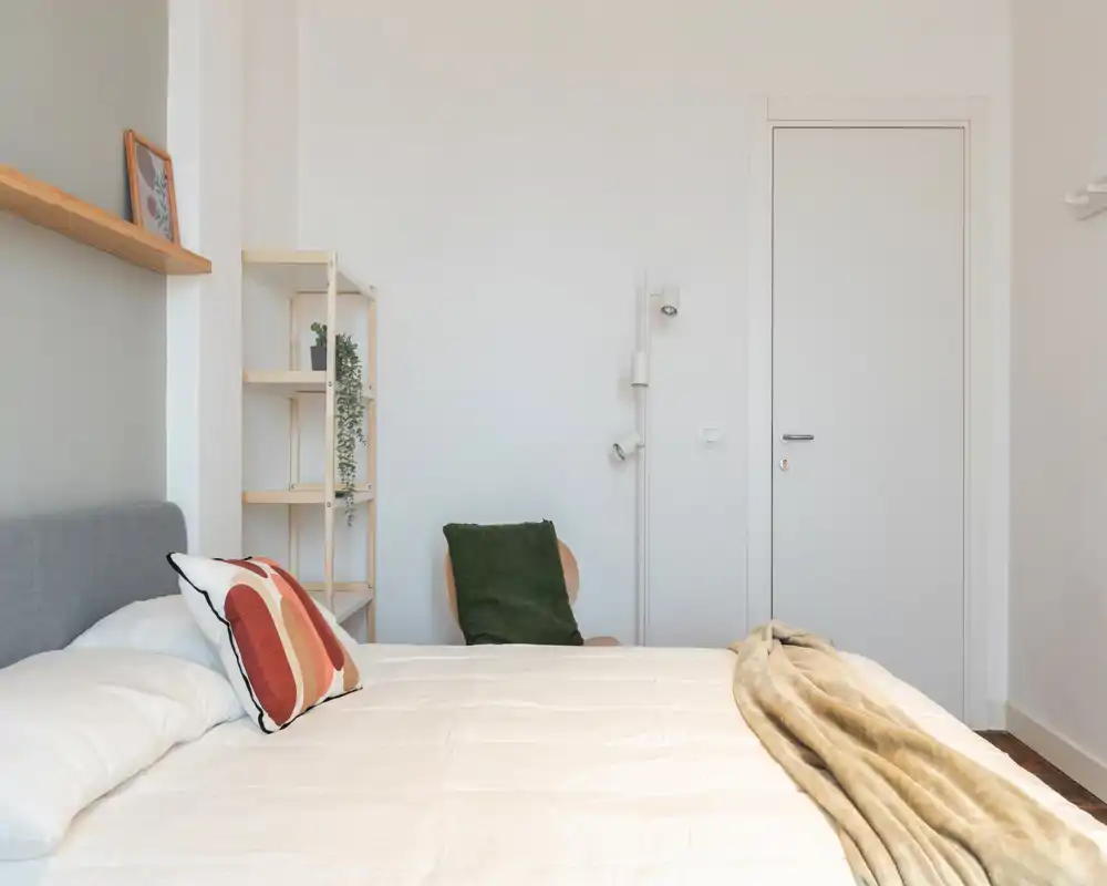 Dormitorio luminoso y minimalista con una gran cama blanca, manta y cojines decorativos, una pequeña estantería y una silla sencilla junto a la pared — ambiente tranquilo y aireado.