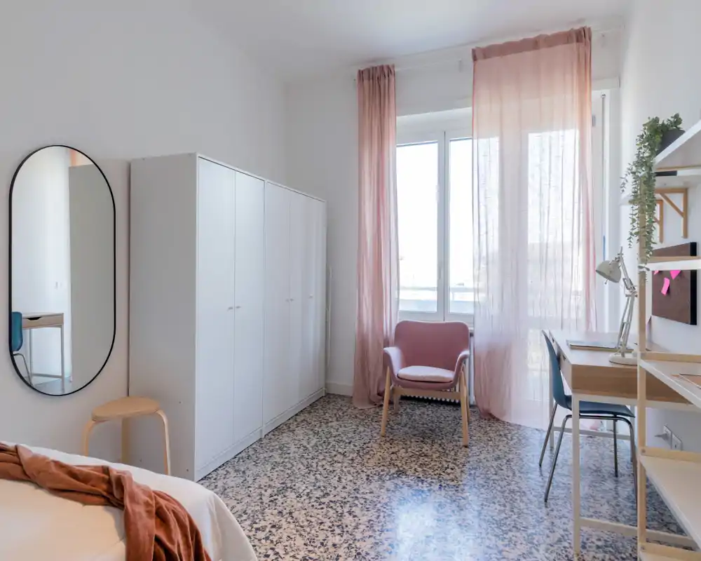 Sunny, stylish bedroom with a wardrobe, desk and a comfortable armchair beside a large window dressed with sheer pink curtains. The bed is partially visible with warm textiles, and the room features terrazzo flooring and bright natural light.