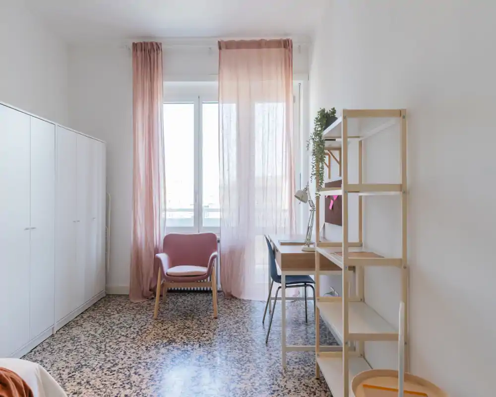 Bright, minimalist bedroom corner with a pink armchair, sheer curtains and a small work desk next to shelving — ideal for a single occupant seeking a calm, well-lit space.