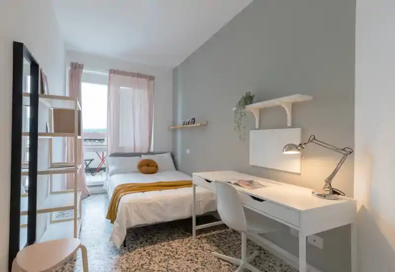 Bright, stylish bedroom with a comfortable double bed, desk workspace and small balcony visible through sheer curtains — ideal for a student or professional.