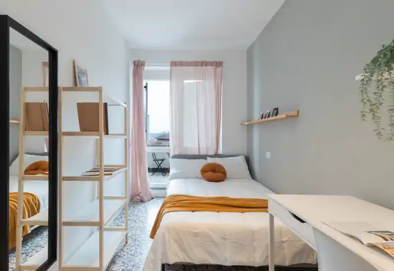 Bright, neatly styled single bedroom with a white bed, warm ochre throw and round cushion; window with soft pink curtains opens to a small balcony, plus a desk and shelving for storage.