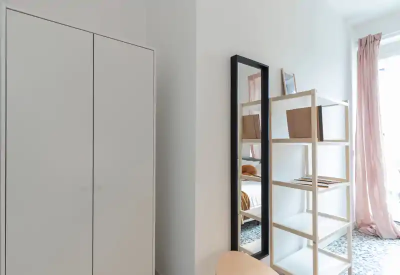 Bright, minimalist bedroom corner with a tall mirror, shelving and a glimpse of the bed. Soft natural light and pale pink curtains give a calm, airy feel.