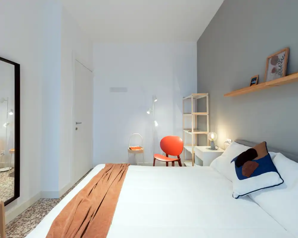 Bright, modern bedroom with a large white bed, decorative pillows and a rust-coloured runner; cozy accents include a red chair, bedside lamp and wall shelf with art.