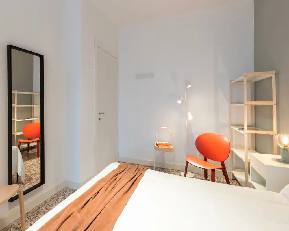 Bright, modern bedroom with a neatly made bed, warm brown throw and an orange accent chair. The room features clean lines, a standing lamp and an open shelving unit, ideal for a cozy, stylish stay.