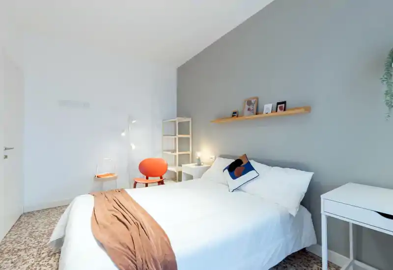 Bright, modern bedroom with a neatly made double bed, soft white linens, decorative cushion and a warm throw; minimalist shelving and a small desk complete the comfortable, well-lit space.