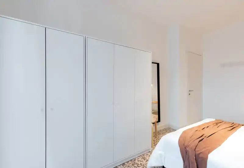 Bright, minimally styled bedroom with a white wardrobe and a neatly made bed featuring a warm brown throw. The room feels clean and airy with patterned flooring and a full-length mirror adding depth.