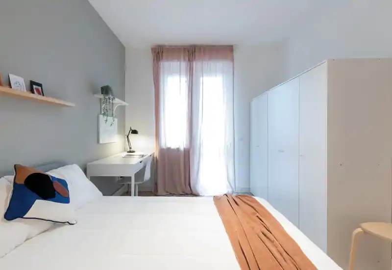 Bright, modern bedroom with a neatly made double bed, soft textiles and a minimalist study desk beside a sunlit window.