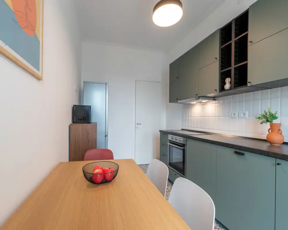 Modern compact kitchen with matte green cabinets, integrated oven and a wooden dining table with chairs; bright, well-styled space ideal for short-term rentals.