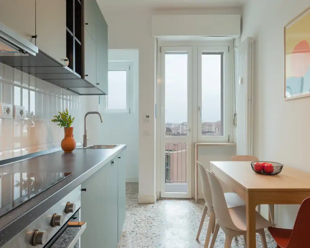 Modern, bright kitchen with built-in oven, induction hob, sink and a small dining table beside a balcony door letting in plenty of natural light.