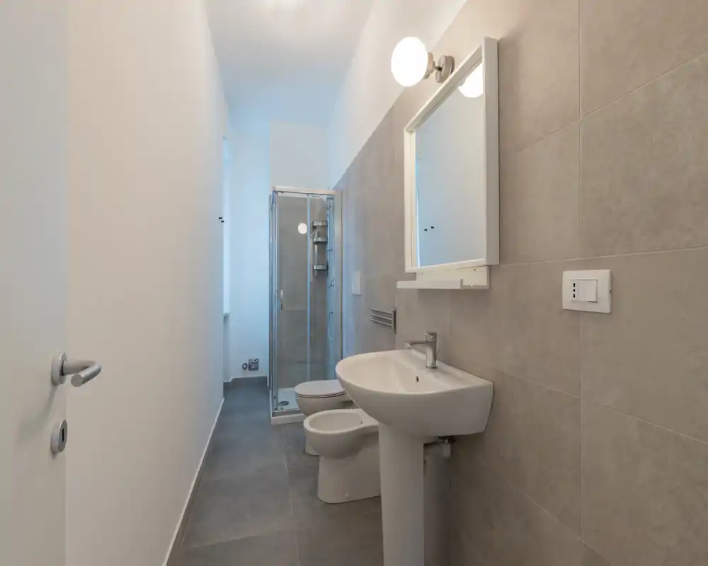 Bright, modern bathroom with a pedestal sink, bidet, toilet and a glass-enclosed shower; neutral gray tiles and a wall-mounted mirror create a clean, minimalist look.