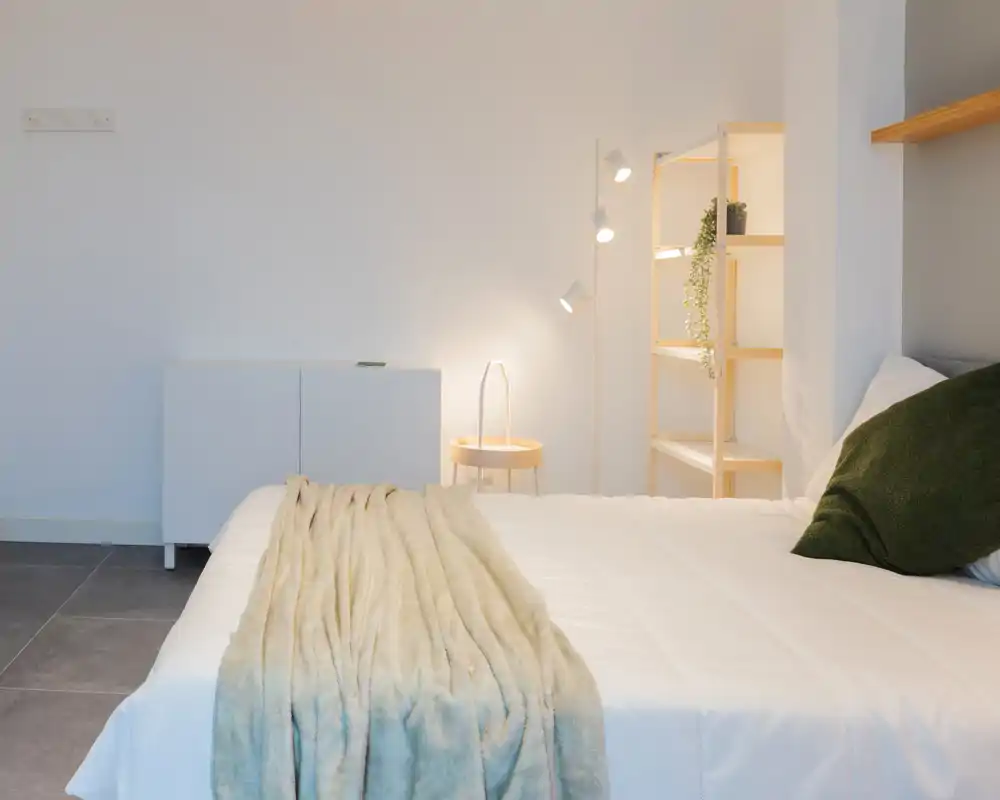 Bright, minimal bedroom with a neatly made bed, soft throw and a cozy green cushion by a light wooden shelving unit.