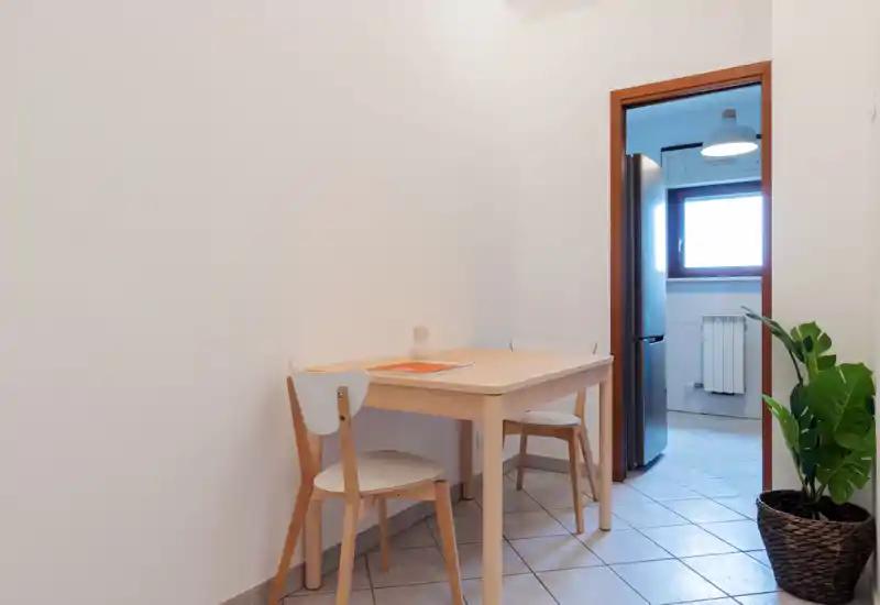 Compact dining nook with a light wood table and two chairs, tiled floor and a potted plant; bright natural light from the adjacent kitchen window.