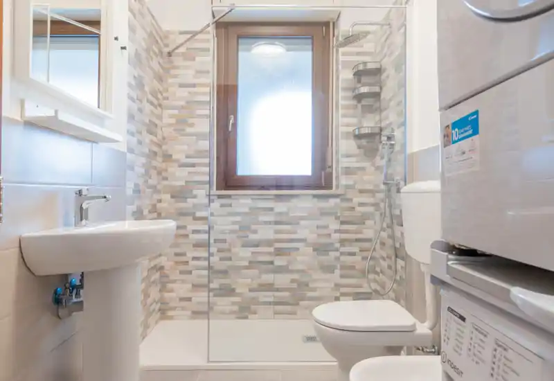 Bright, modern bathroom with a walk-in shower, pedestal sink and toilet; patterned tile wall and frosted window provide privacy.