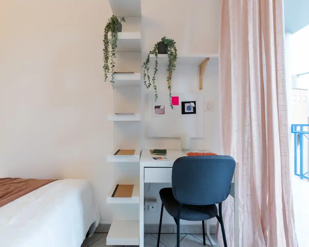 Coin de chambre lumineux et moderne avec un lit bien fait, un petit bureau et des étagères garnies de plantes suspendues. La lumière naturelle provenant du balcon met en valeur les textiles doux et la décoration minimaliste, créant un espace agréable pour dormir et travailler.