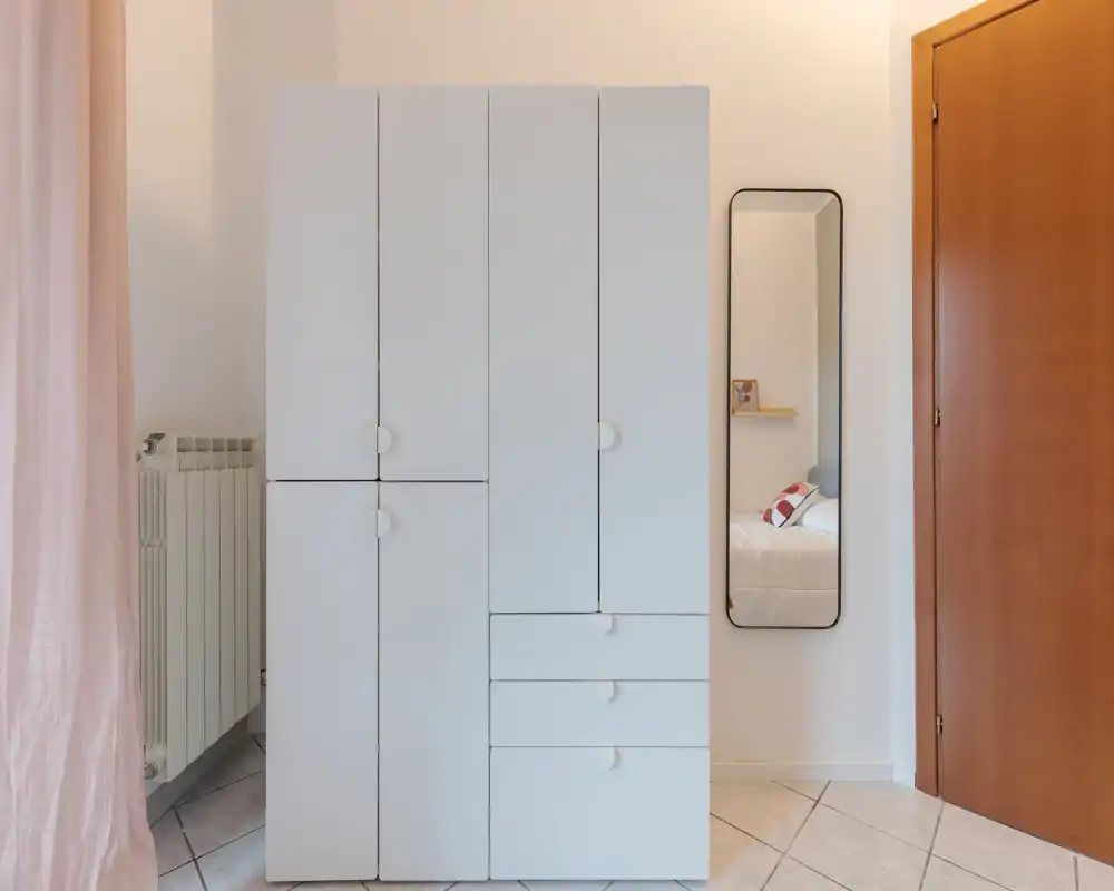 Coin de chambre soigné encadré par une armoire blanche et une fine miroir mural qui reflète partiellement le lit. Lumière naturelle douce et tons pâles pour une ambiance calme et minimaliste.
