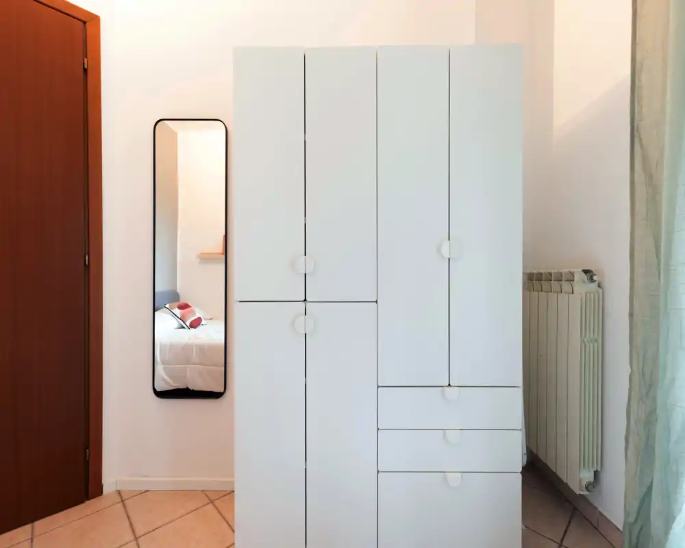 A clean, minimal bedroom view focused on a large white wardrobe with a full-length mirror reflecting a bed with pillows; soft natural light creates a calm atmosphere.