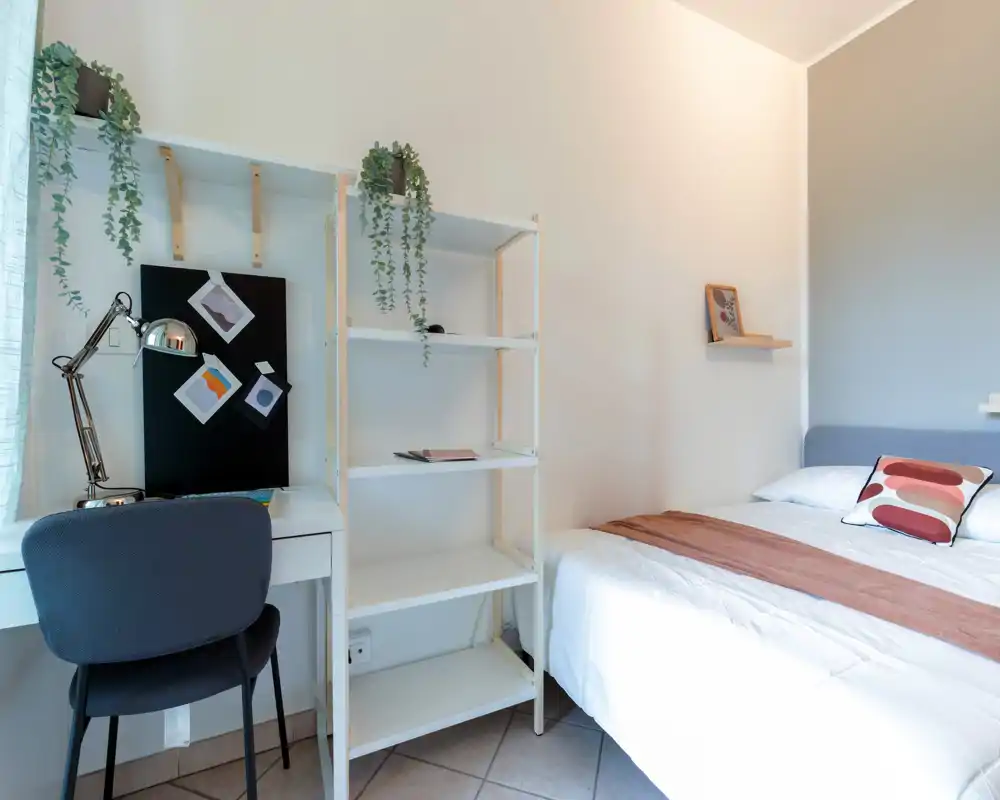 Bright, modern single bedroom with a neatly made bed, bedside shelf and a small workspace with desk and chair — ideal for a student or solo traveler.