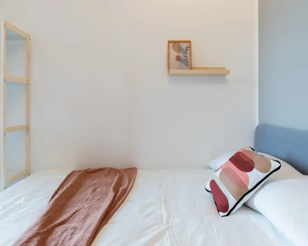 Bright, minimal bedroom with a neatly made bed, decorative cushion and a warm throw; clean white walls and simple shelving create a calm, contemporary feel.