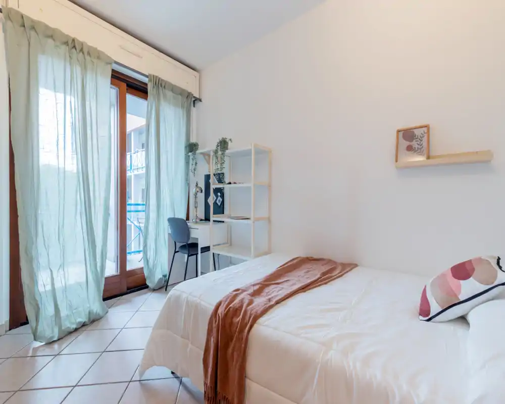 Bright, airy bedroom with a neatly made double bed, soft textiles and a small desk by the balcony providing natural light—ideal for a cozy single or couple stay.